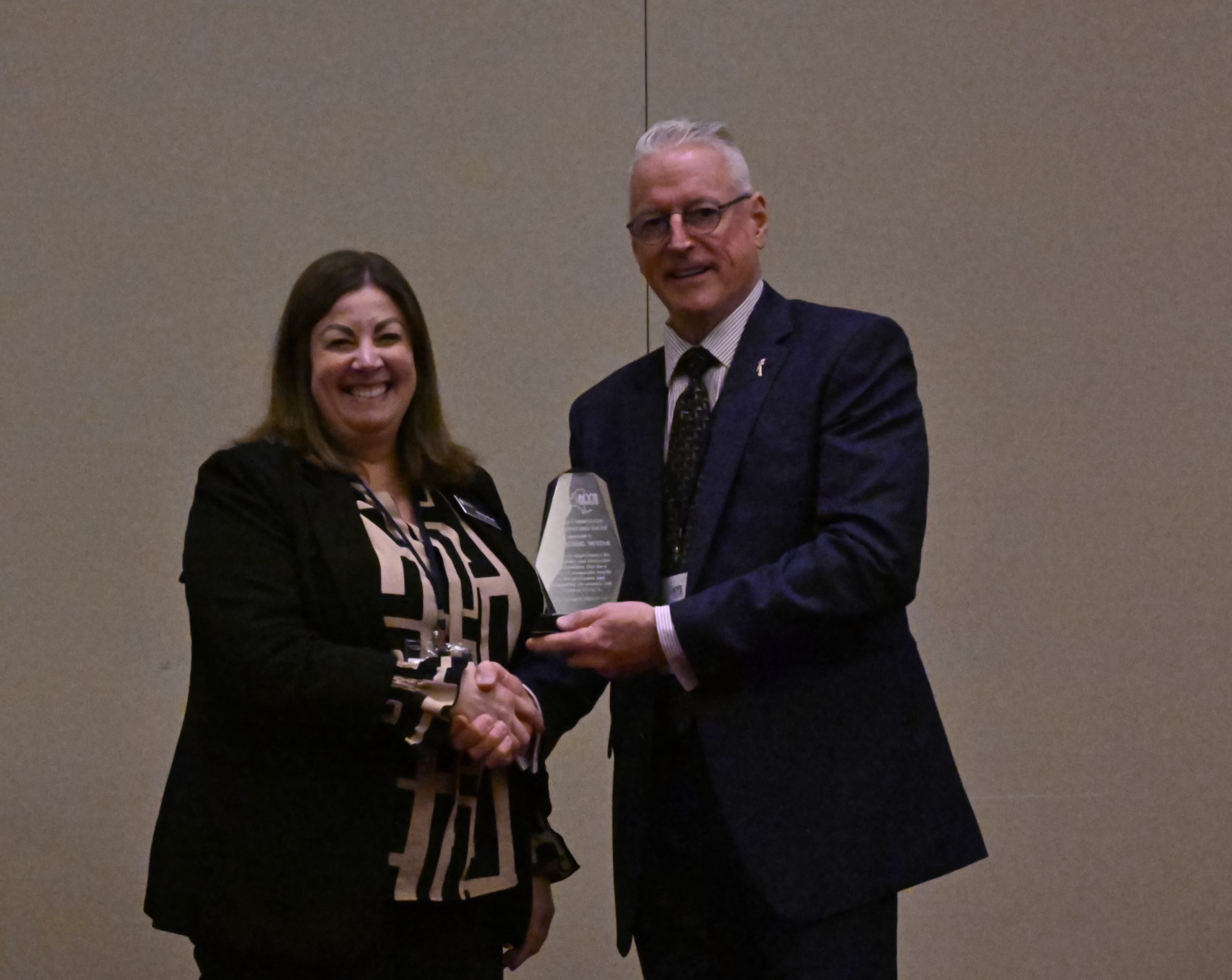Two professionals shake hands as a woman smiles, holding a glass award at a formal event.