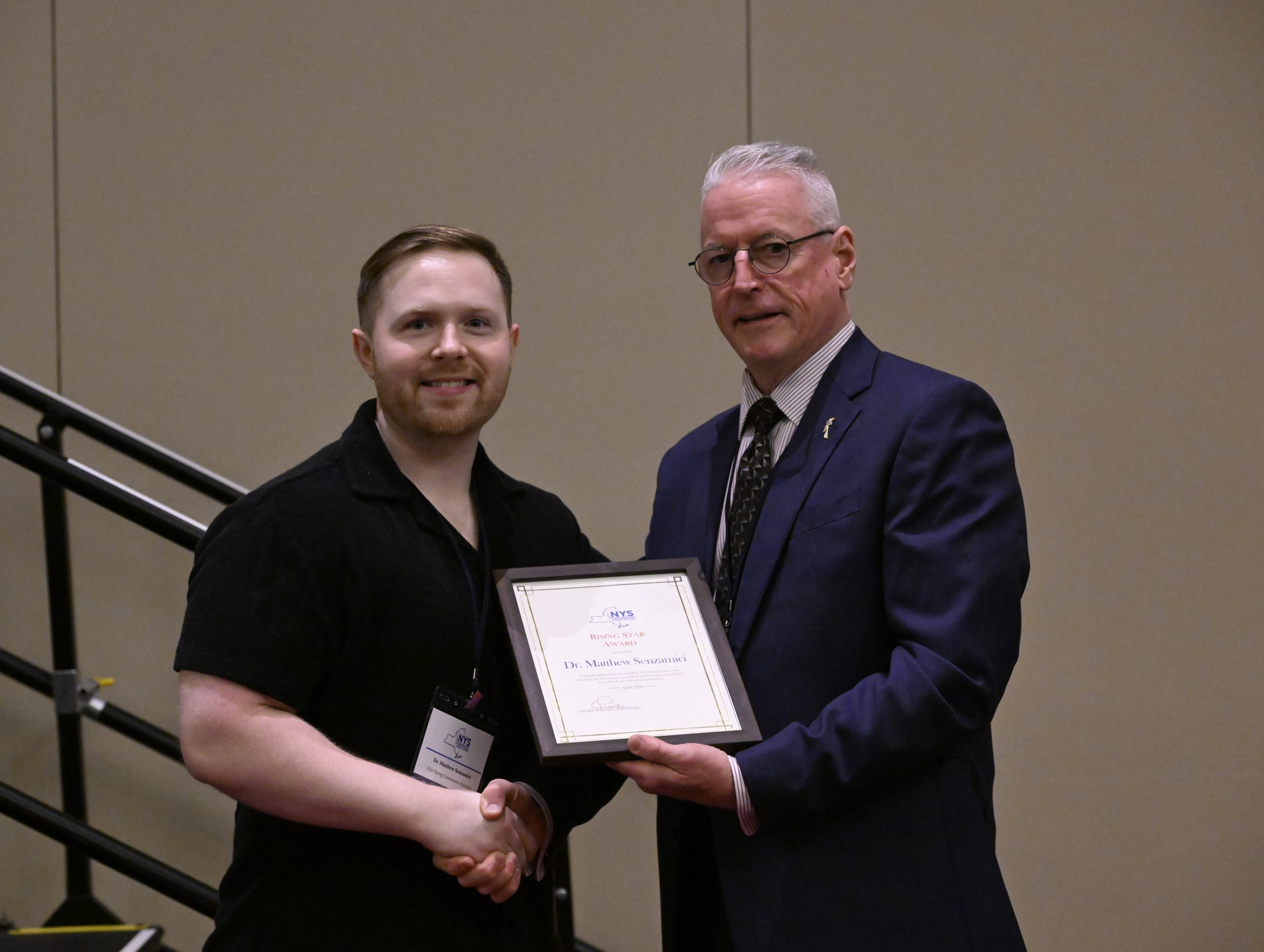 Two men shaking hands as one presents a framed award certificate to the other at a formal event.