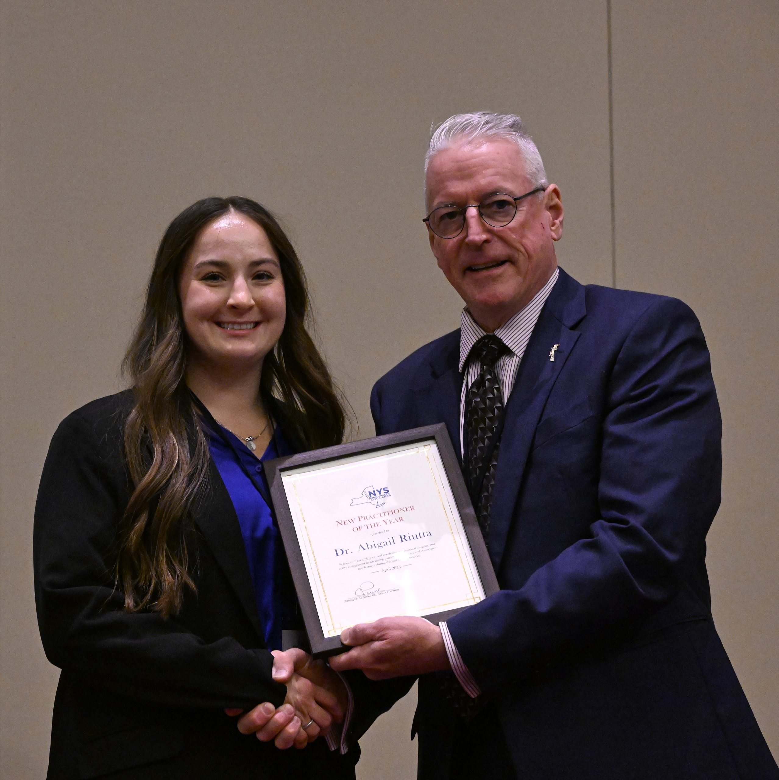 Two professionals in formal attire shake hands; a framed certificate is visible in the foreground.
