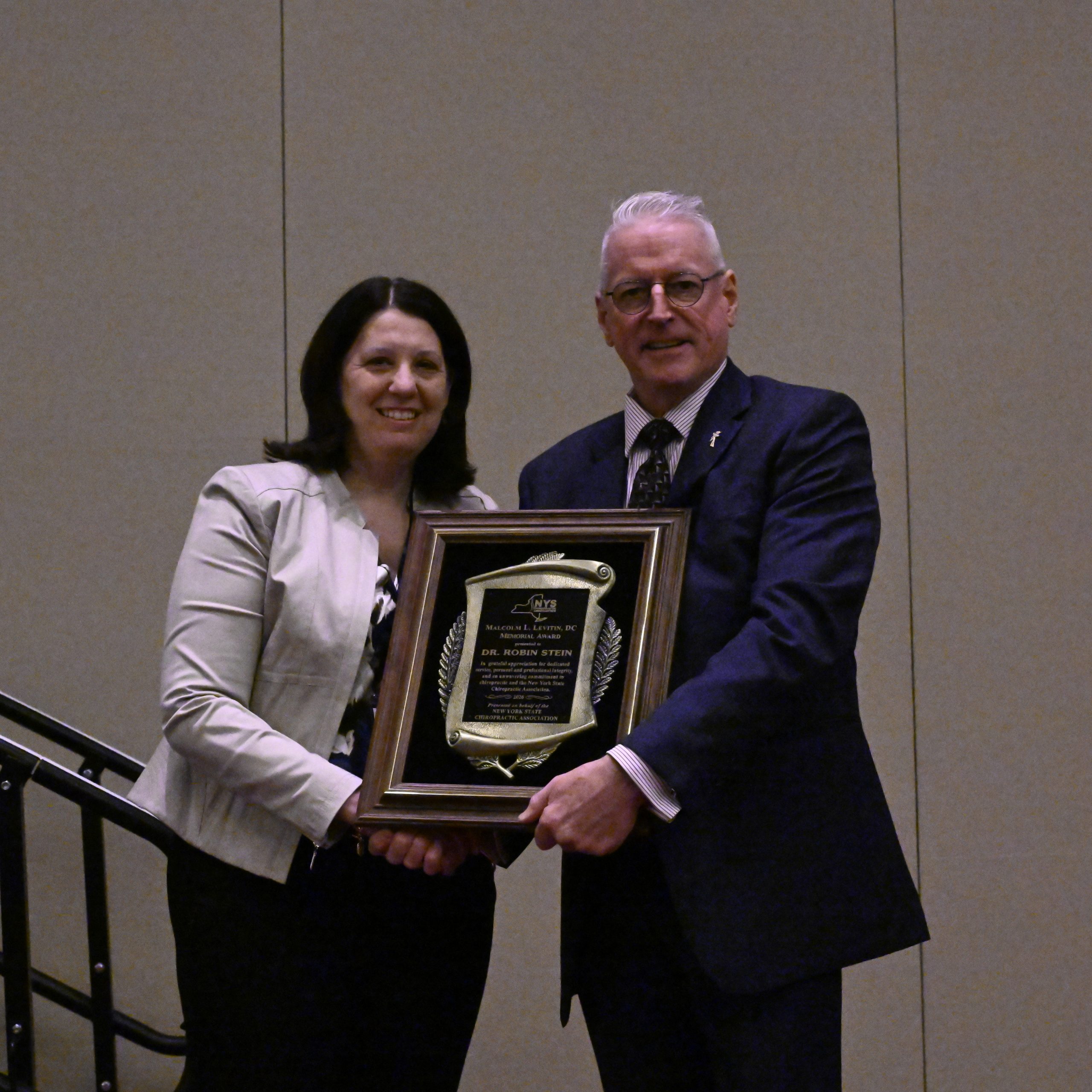 Two people, a woman and a man in a suit, smile while holding a large framed award plaque between them.