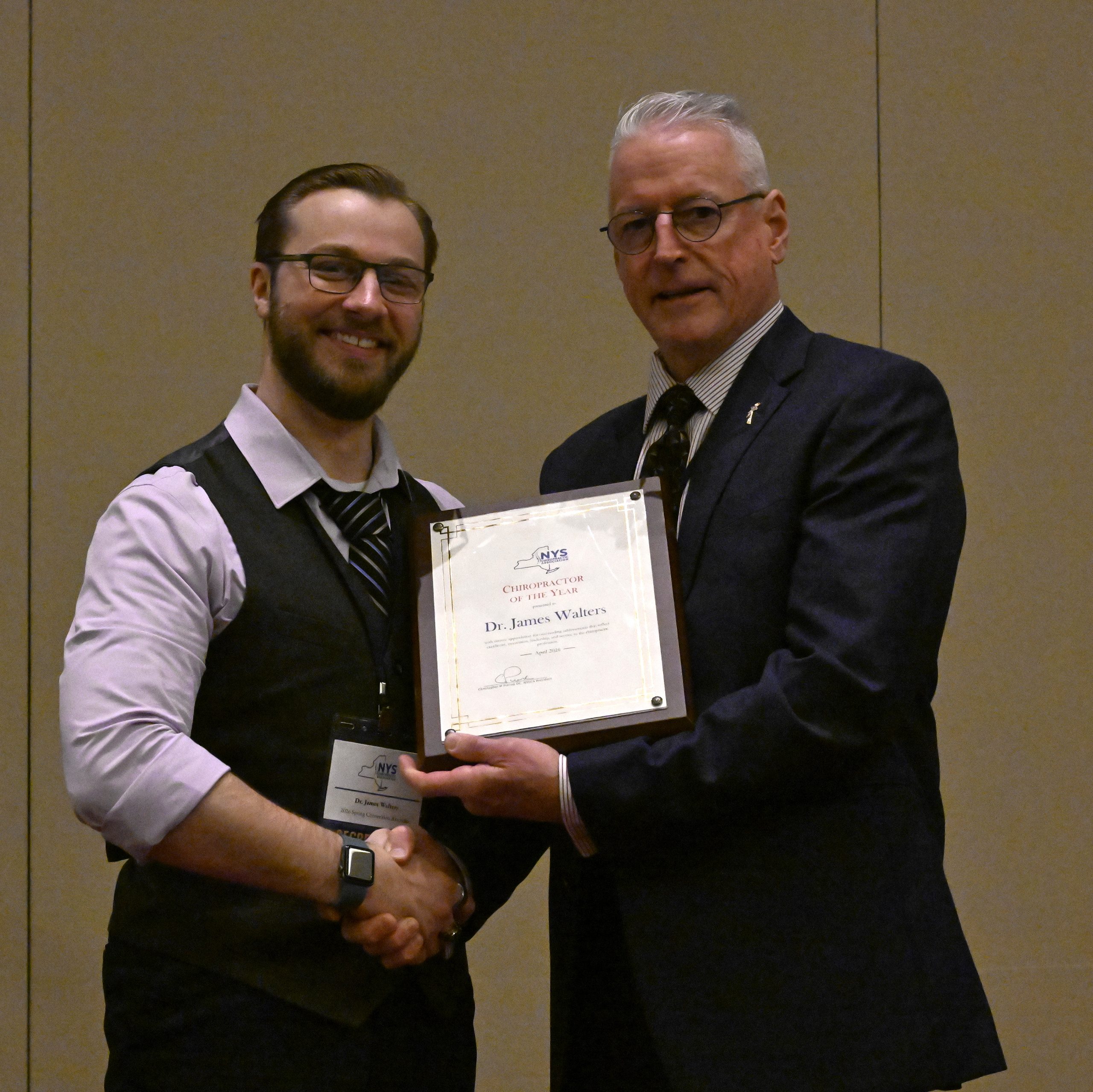 Two men in suits shaking hands at a formal event, one presenting a framed certificate plaque.